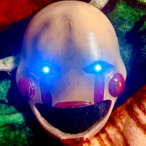 May include: A white mask with blue glowing eyes, red cheeks, and a red mouth. The mask has vertical purple stripes under the eyes and is designed to resemble a puppet or doll.