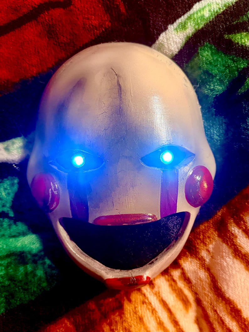May include: A white mask with blue glowing eyes, red cheeks, and a red mouth. The mask has vertical purple stripes under the eyes and is designed to resemble a puppet or doll.