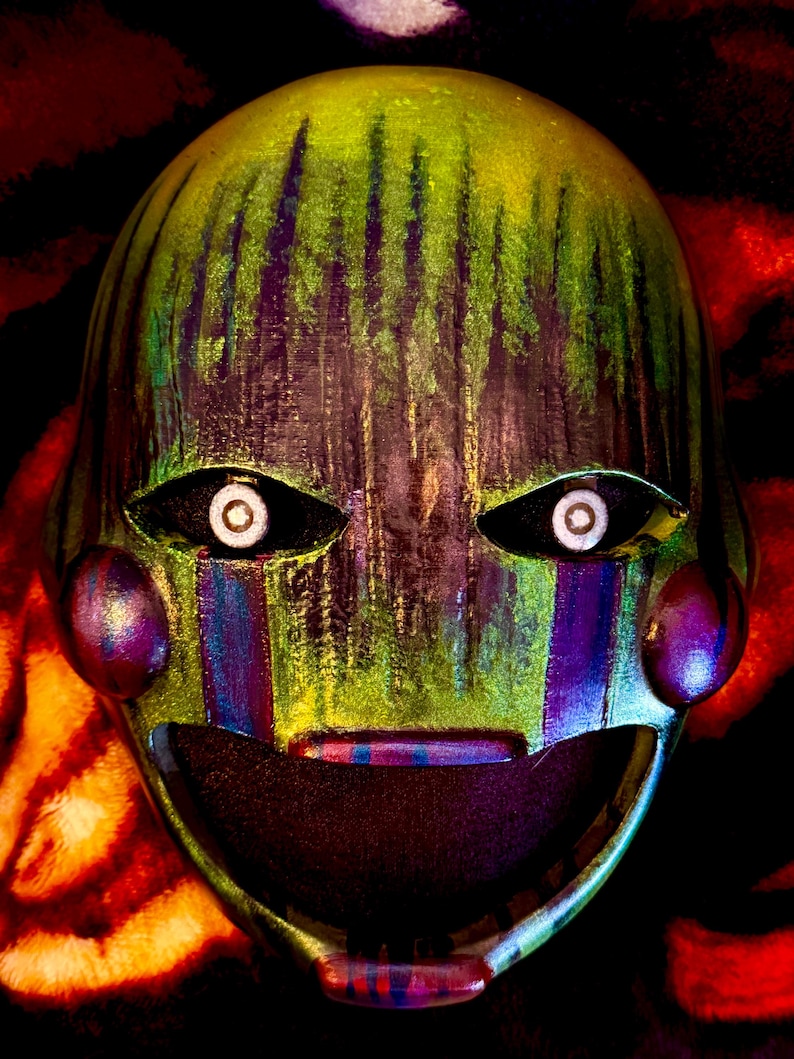 May include: A close-up of a mask with a green and yellow painted surface. The mask has black eye sockets with white circles, purple cheeks, and a wide open mouth. The mask has vertical green and purple stripes on the forehead and cheeks.