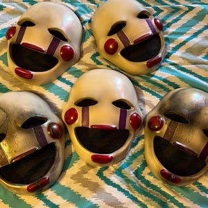 May include: Five white masks with red and purple accents. Each mask has a wide, smiling mouth, red cheeks, and purple tear-like markings under the eyes. The masks are arranged on a blue and white patterned surface.