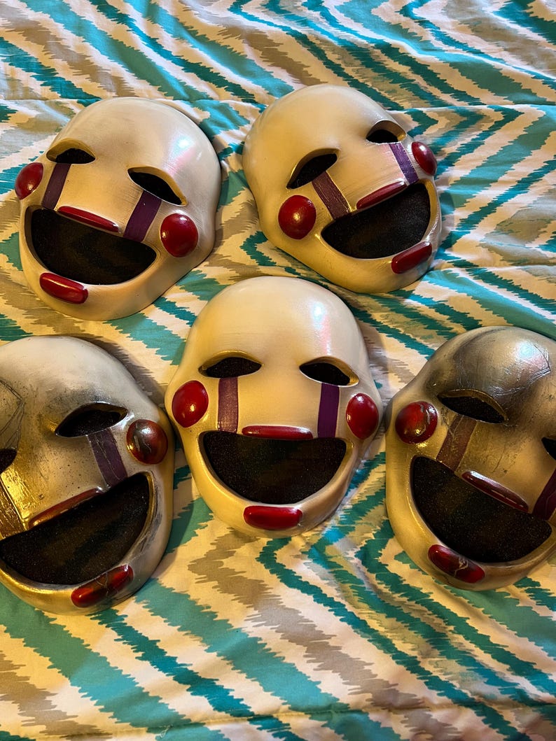May include: Five white masks with red and purple accents. Each mask has a wide, smiling mouth, red cheeks, and purple tear-like markings under the eyes. The masks are arranged on a blue and white patterned surface.