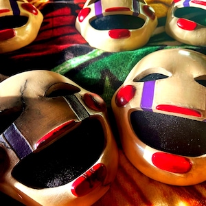 May include: Several beige-colored puppet masks with red lips, cheeks, and accents. The masks have black eye holes and purple stripes on the eyes. The masks are arranged on a colorful, patterned fabric.