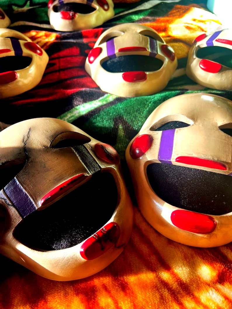 May include: Several beige-colored puppet masks with red lips, cheeks, and accents. The masks have black eye holes and purple stripes on the eyes. The masks are arranged on a colorful, patterned fabric.