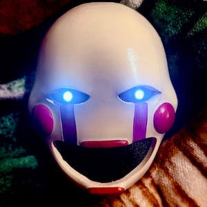 May include: A white mask with glowing blue eyes, red cheeks, and purple tear streaks. The mask has a wide, open mouth with a red painted lip. The mask is likely for cosplay or costume use.