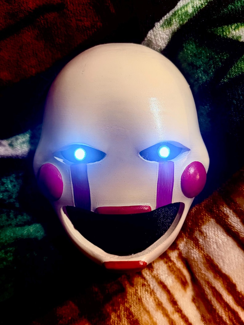 May include: A white mask with glowing blue eyes, red cheeks, and purple tear streaks. The mask has a wide, open mouth with a red painted lip. The mask is likely for cosplay or costume use.