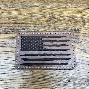 May include: A brown leather patch with a distressed American flag design. The flag is black and white with a white background and black stars and stripes.