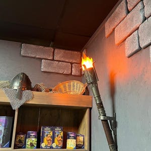 May include: A wall-mounted decorative torch with a flickering flame. The torch has a metal head and a brown handle, mounted on a grey wall. A wooden shelf displays a helmet, chainmail, and various boxes.
