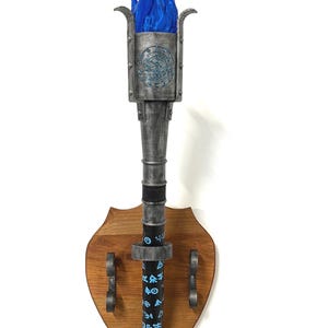Peut inclure: Une torche décorative murale avec un effet de flamme bleue. La torche a une finition métallique de couleur argentée avec des détails complexes et est montée sur une plaque de bois. La torche présente une base en bois en forme de bouclier avec des accents métalliques décoratifs.