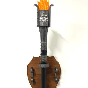 Può includere: Una torcia da parete con una base in legno a forma di scudo. La torcia presenta un design in metallo con un emblema di lupo e una fiamma arancione brillante. La torcia è progettata per essere montata a parete.
