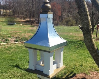 May include: A white and silver birdhouse with a decorative top, mounted on a wooden post. The birdhouse has a classic design with a metal roof and is set in a grassy outdoor area.