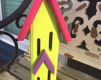 May include: A yellow and pink wooden birdhouse with purple accents. The birdhouse has multiple openings for birds to enter.