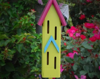 May include: A yellow and pink birdhouse with blue accents. The birdhouse has multiple openings for insects to enter. The birdhouse is mounted on a wooden stake.