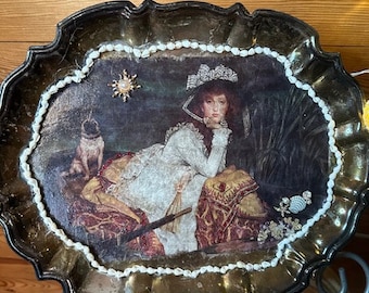 May include: A silver tray with a scalloped edge and a decorative border of white seashells. The tray features a vintage print of a woman in a white dress with a dog, surrounded by flowers and a sun.
