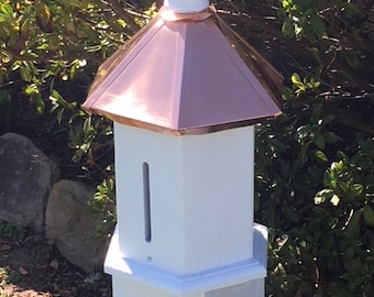 May include: White wooden birdhouse with a copper roof and a white finial. The birdhouse is mounted on a wooden post.