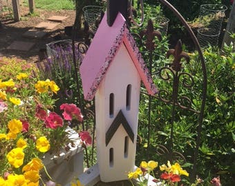 May include: A pink and white birdhouse with a black roof and a black triangular design on the front. The birdhouse is hanging from a metal archway in a garden setting.