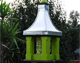 May include: A lime green birdhouse with a silver metal roof and a decorative finial. The birdhouse has a clear glass cylinder inside and is mounted on a black metal post. Red flowers and green foliage are in the background.