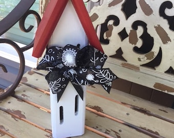 May include: A white wooden house shaped like a church with a red roof and a black and white bow. The house is sitting on a weathered wooden surface.