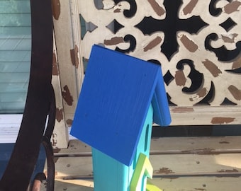 May include: A small blue and turquoise birdhouse with a green perch. The birdhouse has a blue roof and a small opening for birds to enter.