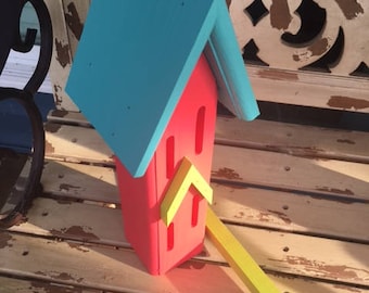 May include: A red birdhouse with a teal roof and a yellow perch. The birdhouse is sitting on a wooden surface.