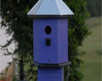 May include: A purple birdhouse with a metal roof, mounted on a black metal stand with decorative scrolls. The birdhouse has a small round entrance hole.