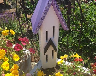 May include: A white birdhouse with a purple roof and black accents. The birdhouse has two openings and is hanging from a black metal hook. The birdhouse is surrounded by yellow and purple flowers.