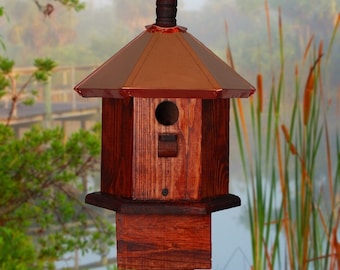 May include: A wooden birdhouse with a copper roof, mounted on a wooden post. The birdhouse has a small opening for birds to enter.