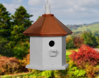 May include: White birdhouse with a copper roof and a small round entrance hole. The birdhouse is hanging from a string and has a decorative finial on top.