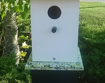 May include: A white birdhouse with a green roof and a black base. The birdhouse has a small hole for birds to enter. The birdhouse is decorated with a floral pattern and a black and white bow.