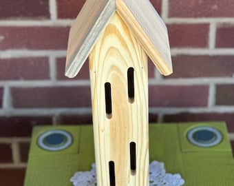 May include: A wooden insect house shaped like a small house with a gray and white weathered roof. The house has multiple openings for insects to enter.