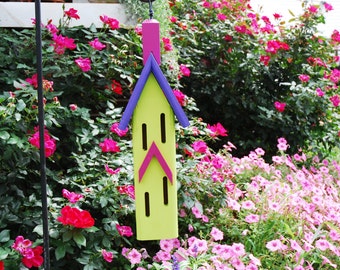 May include: A yellow birdhouse with purple and pink accents is surrounded by pink flowers. The birdhouse has four small windows and a pointed roof.