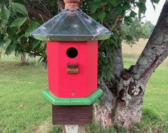 May include: A hexagonal birdhouse featuring a bright red body, a green base, and a metallic silver roof topped with a brown wooden finial. A small circular entrance hole is positioned above a rectangular wooden perch on the front panel.