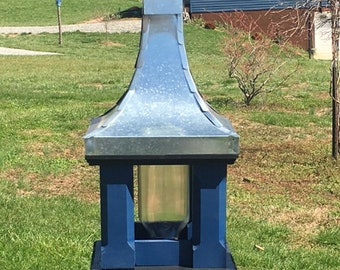 May include: A blue bird feeder with a metal roof and a blue finial. The feeder is mounted on a wooden post and has a clear glass container for birdseed. The feeder is in a grassy yard.