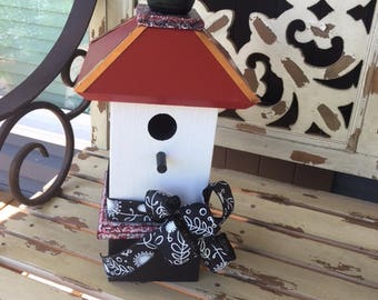 May include: A white birdhouse with a red roof and a black finial. The birdhouse is decorated with a black and white ribbon bow.