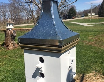May include: A white wooden birdhouse with a galvanized metal roof and a decorative finial. The birdhouse has three entry holes with small perches and is mounted on a wooden post. The birdhouse is outdoors in a grassy area.