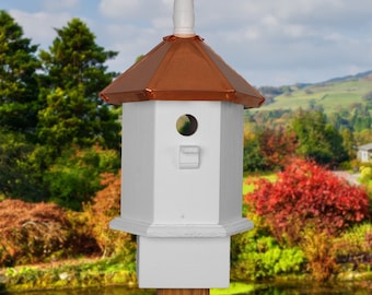 May include: White birdhouse with a copper roof and a small entry hole. The birdhouse is mounted on a wooden post and is surrounded by greenery.