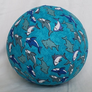 May include: A round, turquoise-colored decorative pillow with a repeating pattern of sharks in various shades of blue, gray, and white. The sharks are stylized with detailed fins and teeth, creating a playful, aquatic theme.