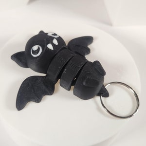 May include: A black bat-shaped keychain with white eyes and a silver ring. The bat is segmented and has a 3D printed look.