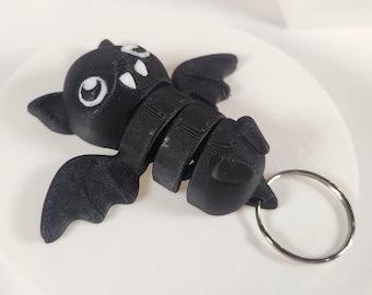 May include: A black bat-shaped keychain with white eyes and a silver ring. The bat is segmented and has a 3D printed look.