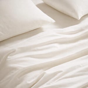 May include: A set of off-white bed linens featuring two pillows and a matching flat sheet. The fabric displays a smooth, soft texture with gentle folds and creases across the surface, creating a clean and neutral appearance for bedding.