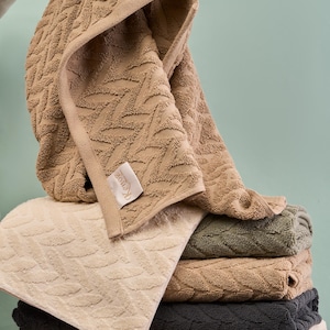 May include: A pile of patterned bath towels in shades of beige, cream, and grey. Each towel has a textured leaf design. The top towel is lifted, showing the brand label. The towels are made of soft material.
