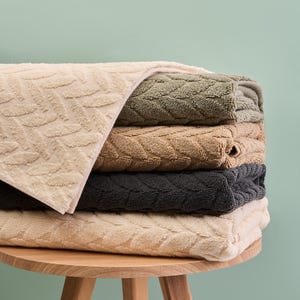 May include: A stack of bath towels in shades of beige, olive green, tan, charcoal grey, and cream. Each towel has a leaf pattern. The towels are stacked on a small, round wooden table.