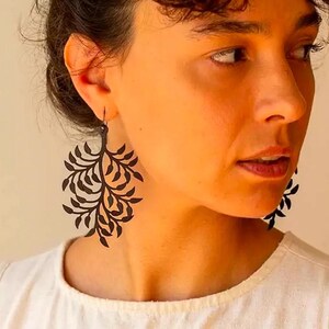 May include: Black, leaf-shaped earrings. The earrings are worn by a person wearing a white shirt. The earrings have a detailed, openwork design of leaves and branches. The earrings are a dark colour, possibly black or a very dark brown.