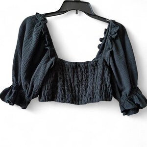 May include: A dark gray crop top with a square neckline and ruffled trim. The top features long sleeves with ruffled cuffs and a smocked bodice. The garment is hanging on a black hanger against a white background.