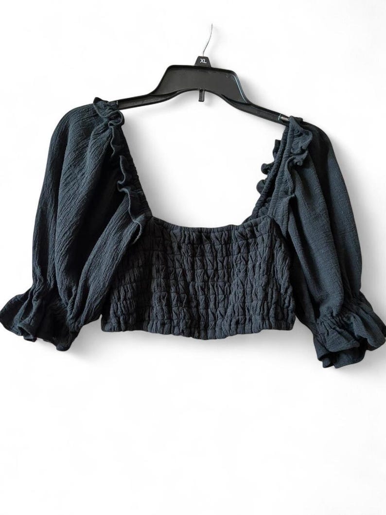 May include: A dark gray crop top with a square neckline and ruffled trim. The top features long sleeves with ruffled cuffs and a smocked bodice. The garment is hanging on a black hanger against a white background.