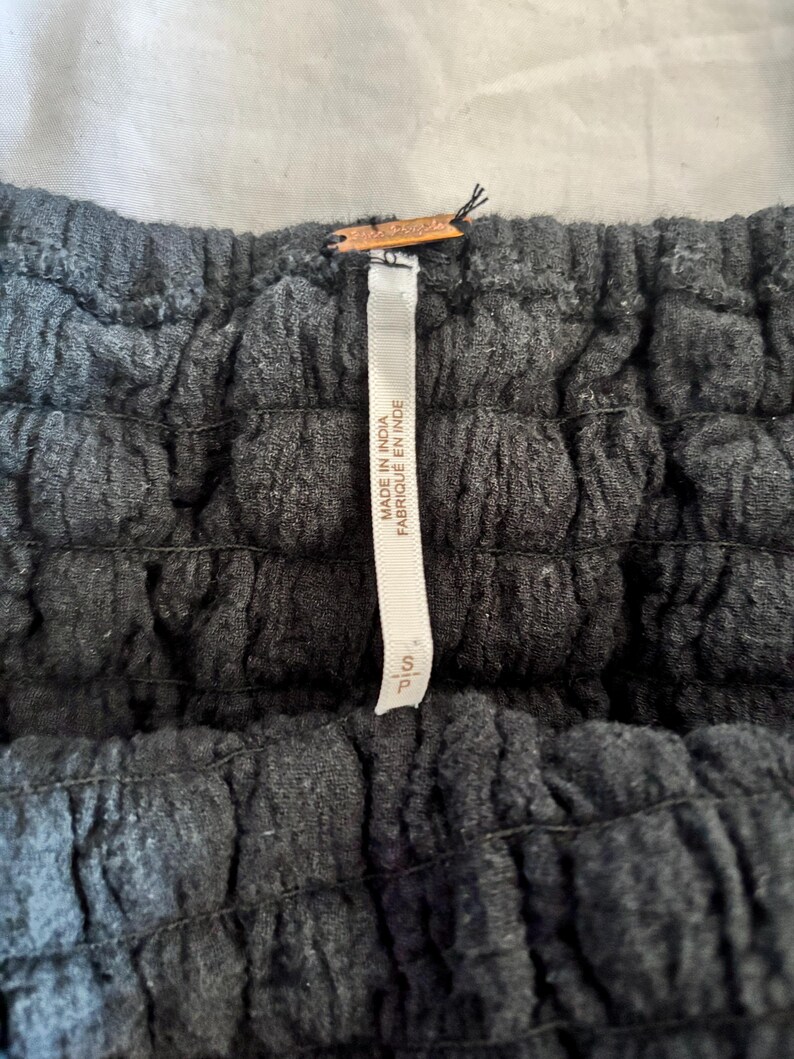 May include: Close-up of a dark gray garment with a textured, ruched design. A white tag with the text "MADE IN INDIA FABRIQU&Eacute; EN INDE" is attached. The tag also has the size "S/P".