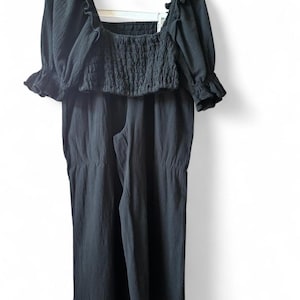 May include: A black two-piece outfit, consisting of a square-neck crop top with ruffled sleeves and a pair of wide-leg pants. The top features a smocked bodice. The outfit is hanging on a black hanger against a white background.
