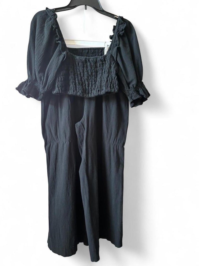 May include: A black two-piece outfit, consisting of a square-neck crop top with ruffled sleeves and a pair of wide-leg pants. The top features a smocked bodice. The outfit is hanging on a black hanger against a white background.