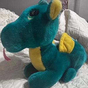 May include: Teal plush dragon toy with yellow accents on the ears and wings. The dragon has a pink tongue and a friendly expression. The stuffed animal is sitting on a white quilted surface.