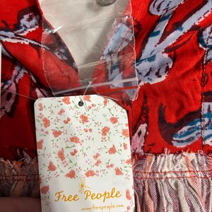 May include: Close-up of a red fabric with a floral pattern in blue and white. The garment has a V-neck and a button closure. A tag with the words "Free People" and a floral design is attached.