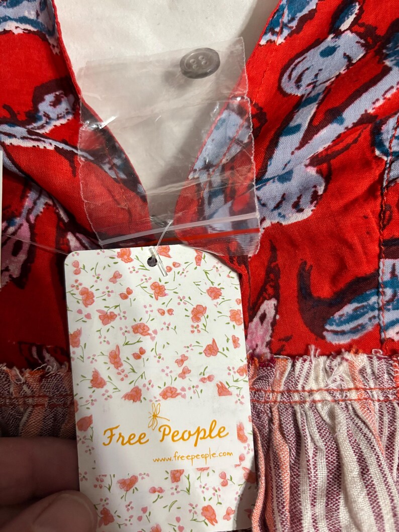 May include: Close-up of a red fabric with a floral pattern in blue and white. The garment has a V-neck and a button closure. A tag with the words "Free People" and a floral design is attached.